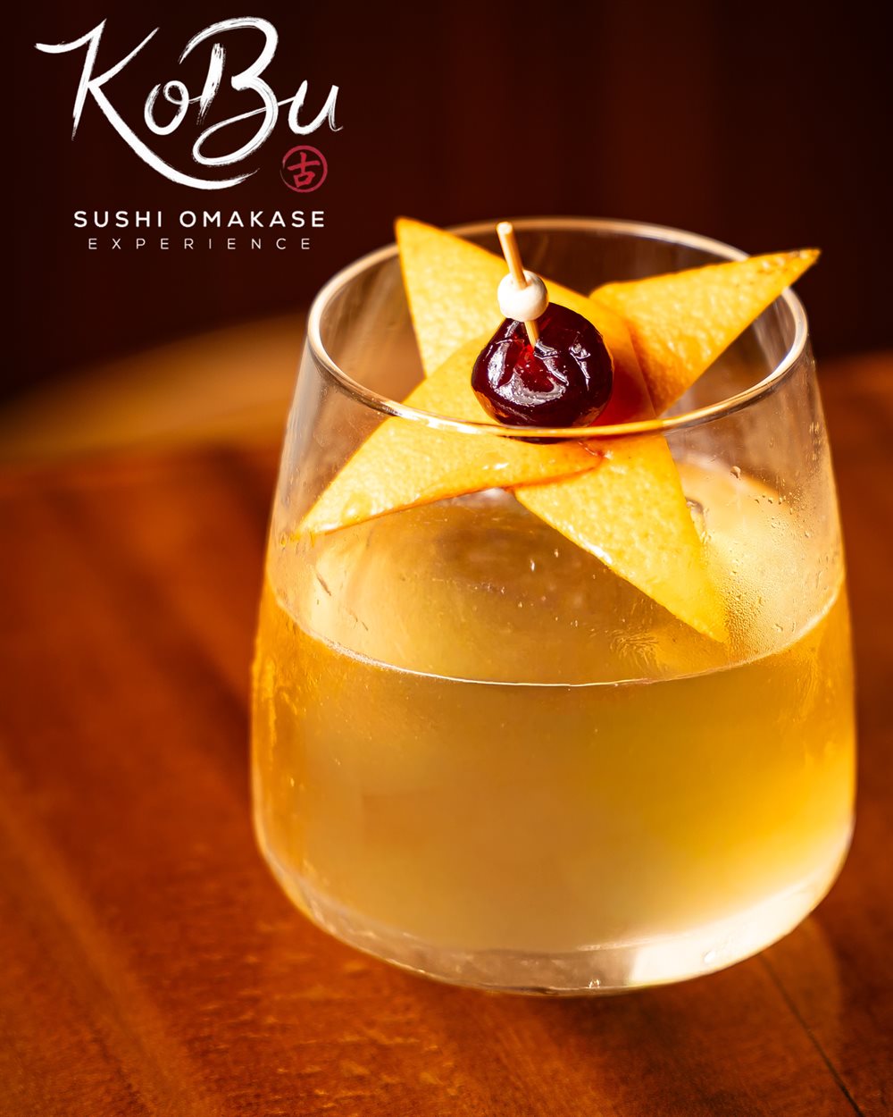 A pale golden cocktail over a clear ice sphere, garnished with a folded citrus peel and a dark cherry on a pick